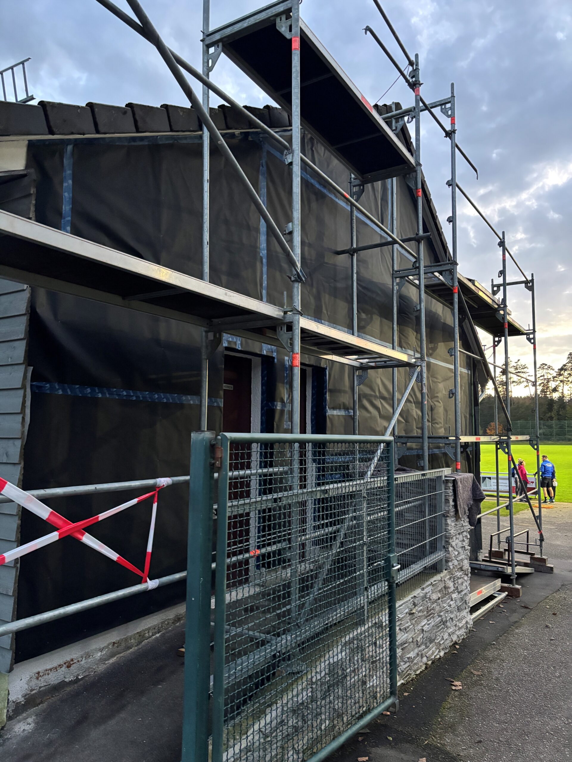 Gerüstbau an einer Fassade eines einstöckigen Hauses mit Spielfeld im Hintergrund