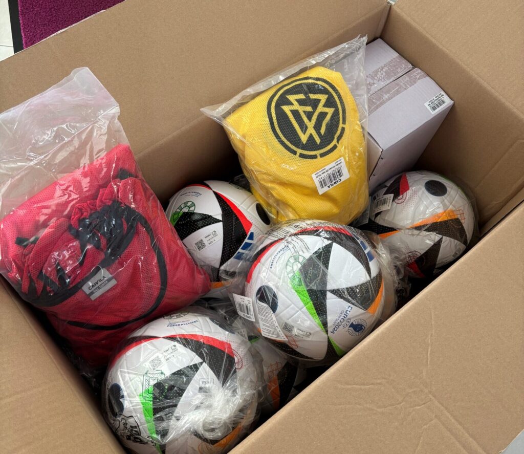 Geöffneter Karton mit mehreren Fußballen und Trainingstrikots in gelb und rot