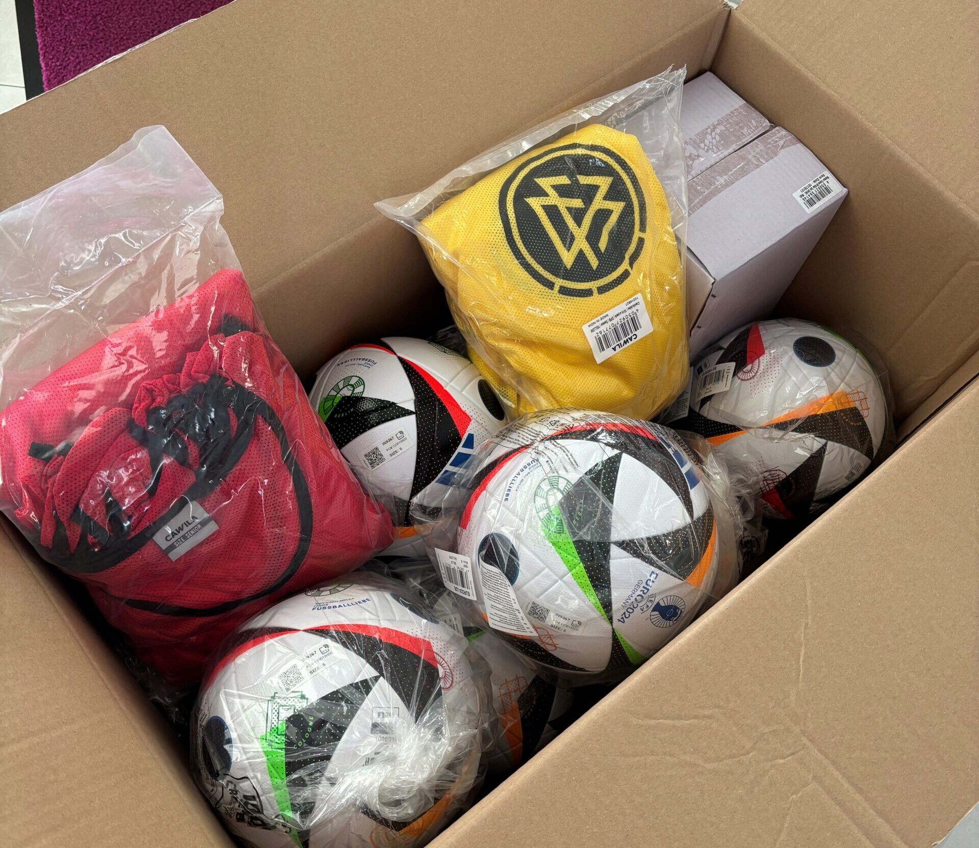 Geöffneter Karton mit mehreren Fußballen und Trainingstrikots in gelb und rot