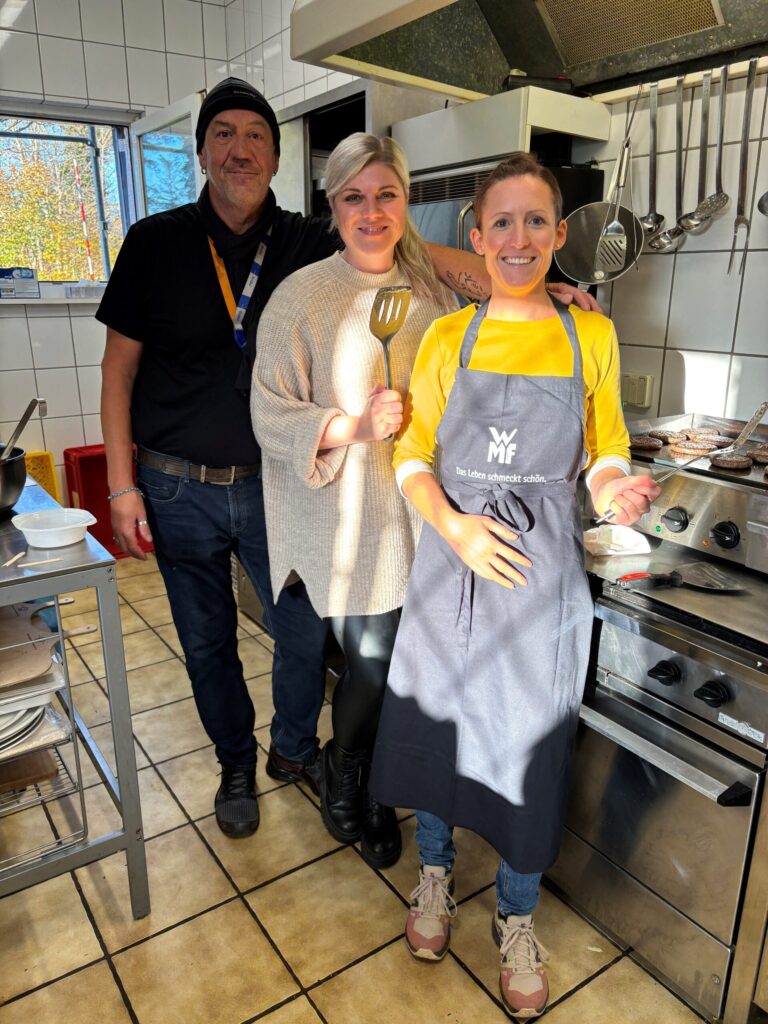 3 Personen nebeneinander in einer Küche posierend und Küchenutensilien hochhebend