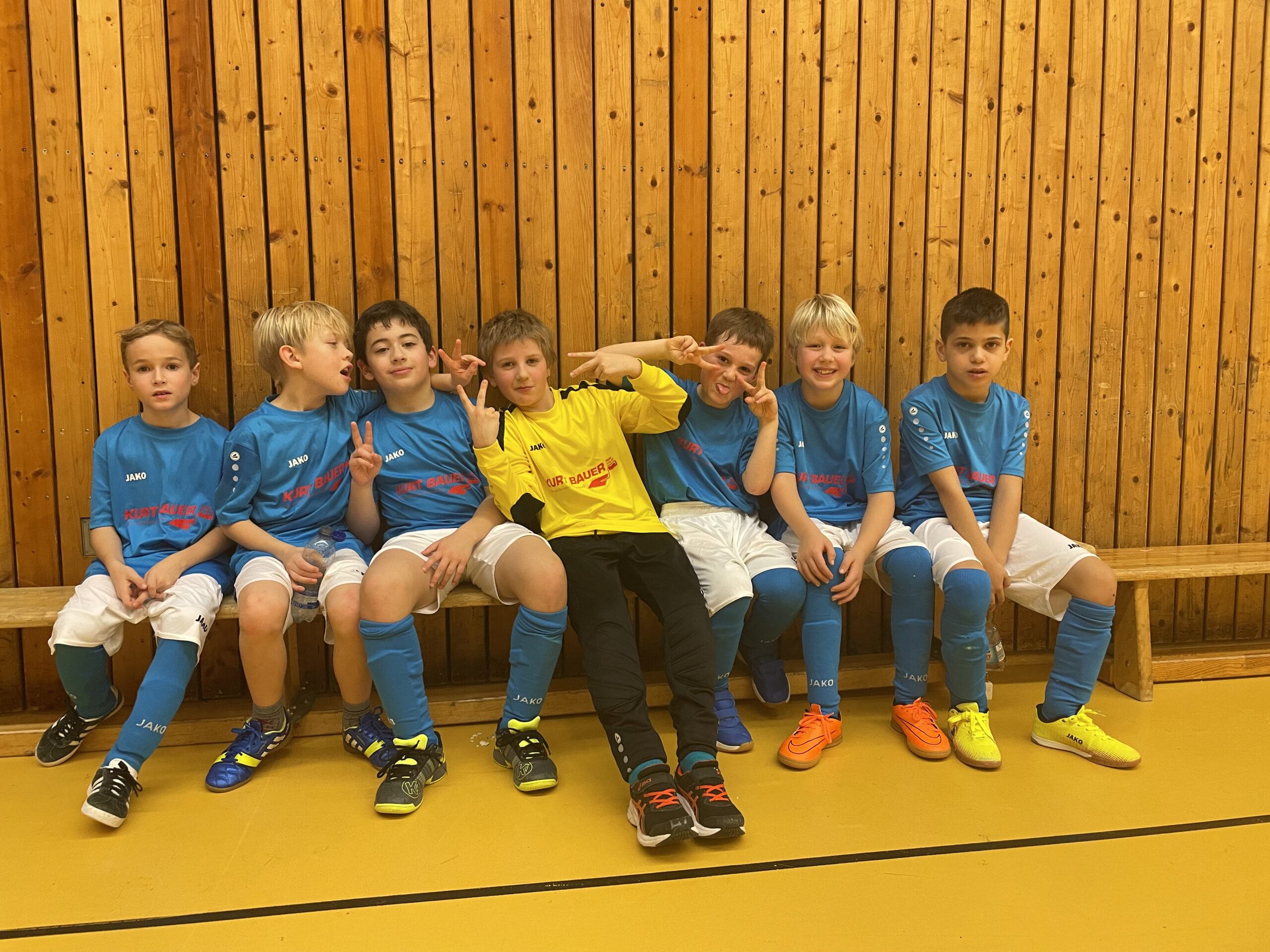 Gruppenbild: 7 jugendliche Fußballspieler und Torwart sitzen in einer Turnhalle auf einer Bank in Fußballtrikots